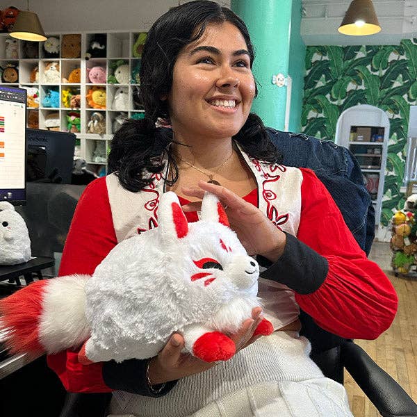 Mini white-and-red kitsune plush held in hands, showing face and red ears.