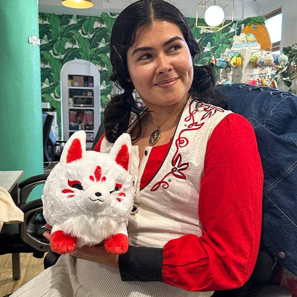 Mini white-and-red kitsune plush held near chest, with red ears and red tail detail.